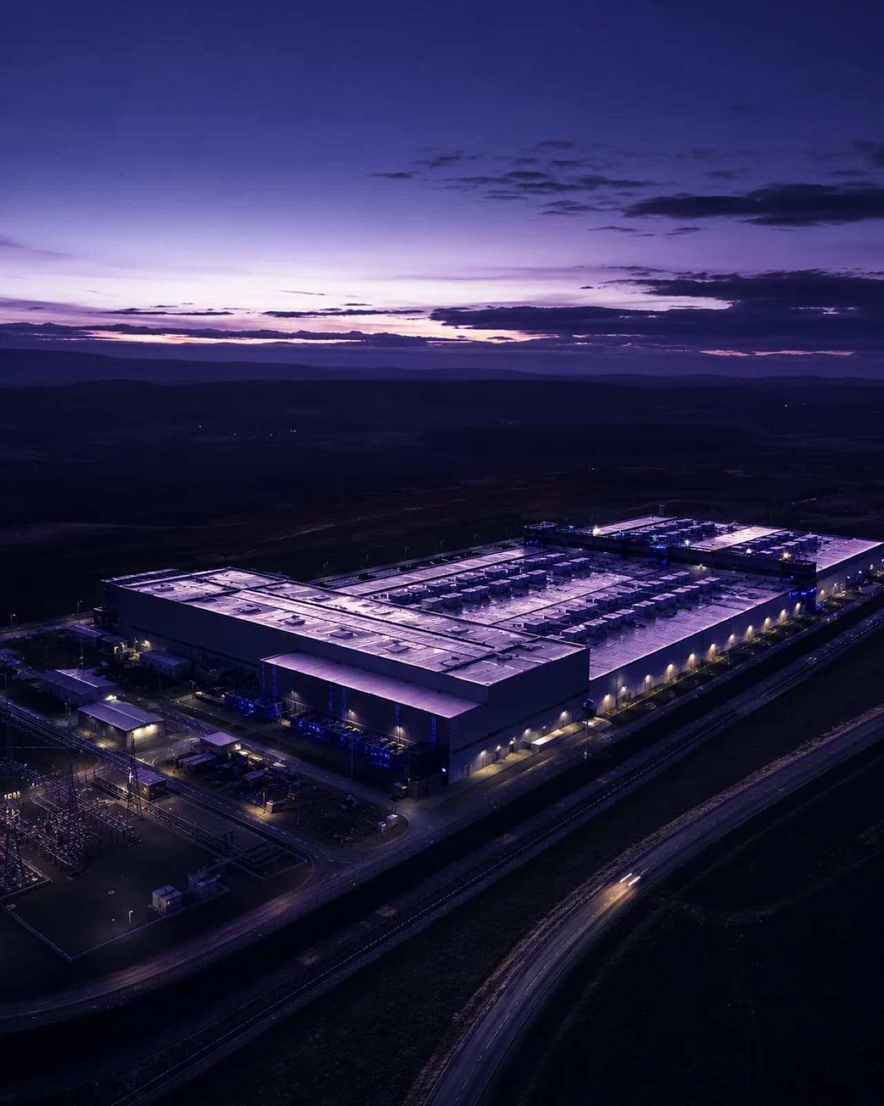 Vista aérea al anochecer de un centro de datos junto a su subestación eléctrica, bajo un cielo violeta con nubes bajas.